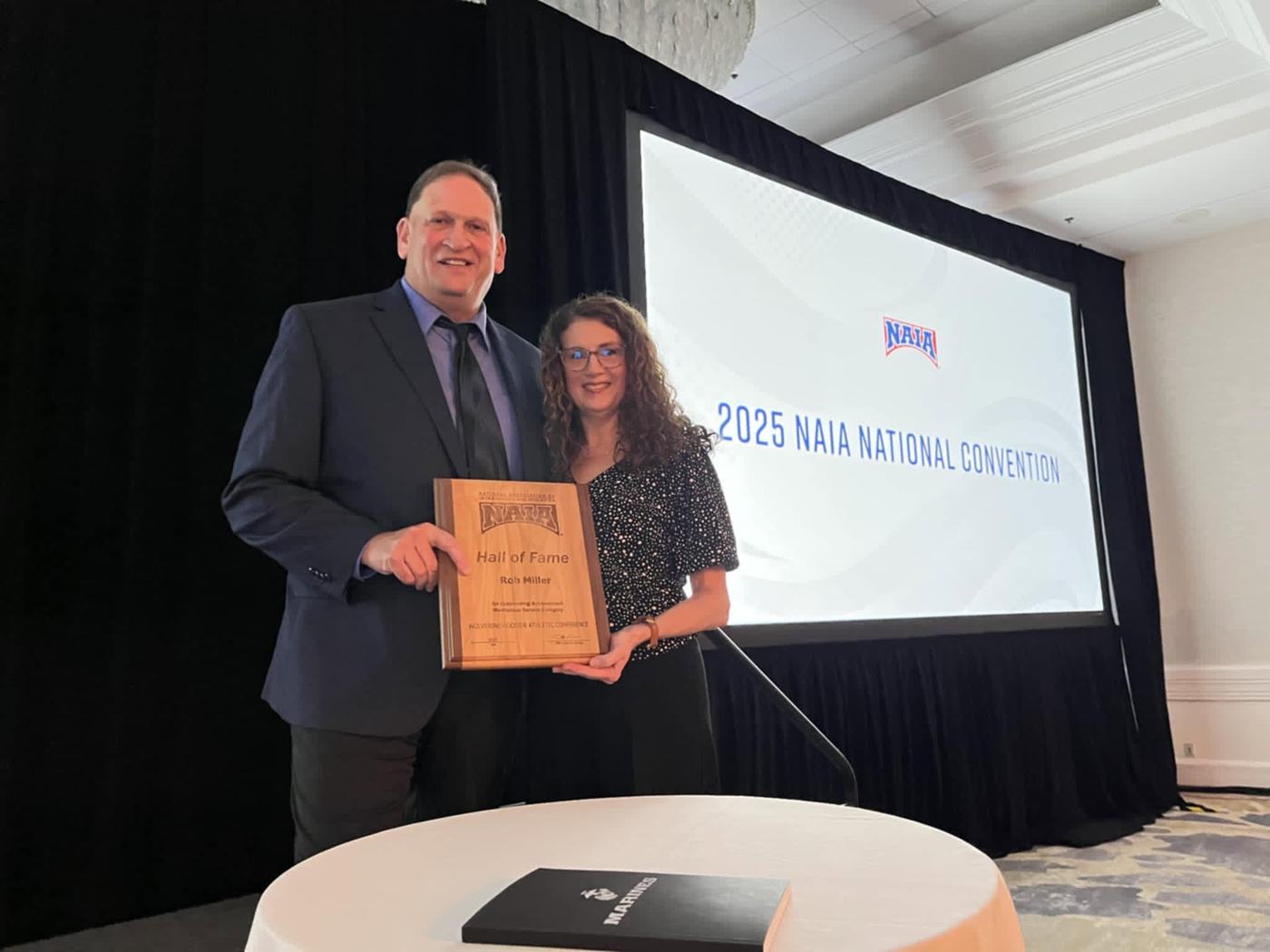Rob Miller and his wife Susan holding his NAIA Hall of Fame plaque on stage at the 2025 NAIA National Convention